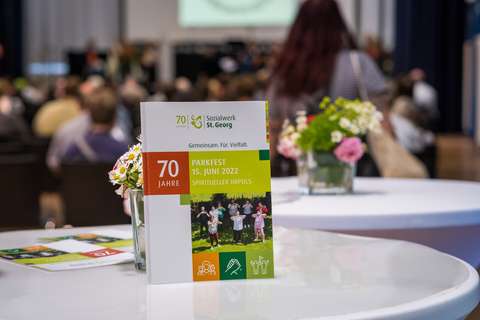 Ein Flyer mit der Aufschrift „70 Jahre Sozialwerk St. Georg Parkfest 15. Juni 2022 Spiritueller Impuls“ steht auf einem runden Tisch, daneben ein Glas mit Blumen; im Hintergrund sitzen mehrere Personen in einem Saal.