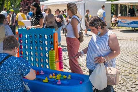 Mehrere Personen spielen an einem blauen Stand mit kleinen Plastikenten und Angeln, im Hintergrund stehen weitere Personen vor weißen Zelten.