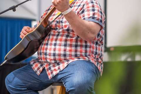 Eine Person mit kariertem Hemd sitzt auf einem Hocker, spielt Gitarre und singt in ein Mikrofon.