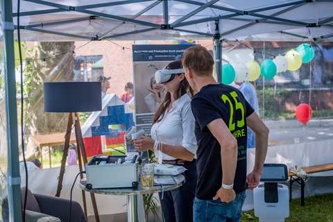 Zwei Personen stehen unter einem Pavillon, eine Person trägt eine VR-Brille und hält Controller, die andere Person steht daneben; im Hintergrund sind bunte Luftballons und Möbel zu sehen.
