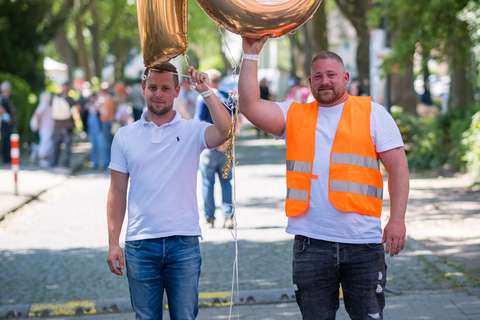 Zwei Personen stehen auf einer Straße, halten goldene Luftballons in Form der Zahl 10, eine Person trägt eine orange Warnweste.