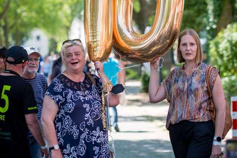 Zwei Personen halten eine große goldene Luftballon-Zahl „10“ im Freien, umgeben von weiteren Menschen und Bäumen.