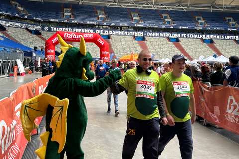 Eine Person im Drachenkostüm gibt einer Person mit Startnummer 3089 die Hand, während mehrere Personen in Sportkleidung an einem Zieleinlauf in einem Stadion vorbeigehen.
