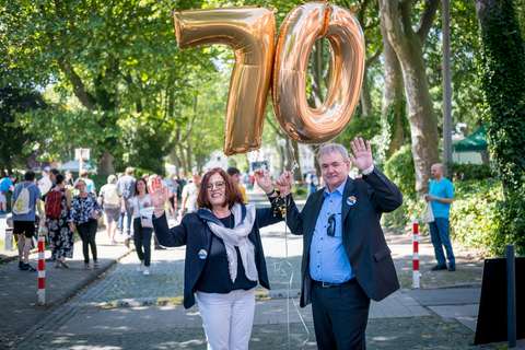 Zwei Personen halten goldene Luftballons in Form der Zahl 70 und winken, im Hintergrund gehen mehrere Personen auf einer von Bäumen gesäumten Straße.
