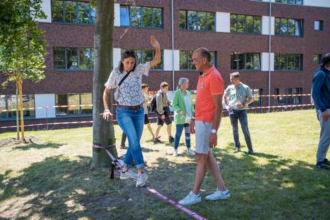 Eine Person balanciert auf einem Slackline-Band zwischen zwei Bäumen, während eine weitere Person daneben steht; im Hintergrund sind mehrere Personen und ein Gebäude zu sehen.