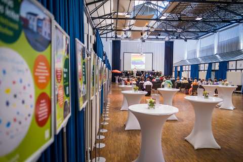 Mehrere Personen sitzen in einem großen Saal vor einer Bühne mit Leinwand, im Vordergrund stehen runde Stehtische mit Blumendekoration und an der linken Seite sind Infotafeln aufgestellt.