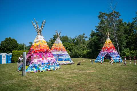 Drei bunte Tipis aus Patchwork-Stoffen stehen auf einer Wiese, eine Person steht neben dem linken Tipi, im Hintergrund sind Bäume und zwei mobile Toiletten.