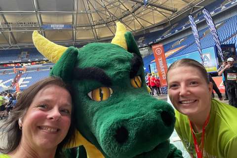 Zwei Personen und ein großes grünes Maskottchen mit gelben Hörnern stehen in einem Stadion.