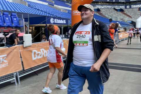 Eine Person mit Startnummer und Kappe geht in einem Stadion unter einem orangefarbenen Zielbogen, im Hintergrund weitere Personen.