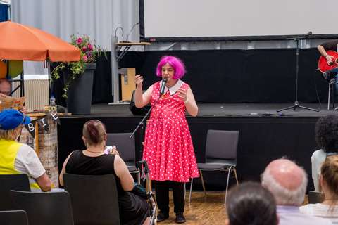Eine Person mit pinker Perücke und gepunktetem Kleid steht an einem Mikrofon vor sitzendem Publikum.