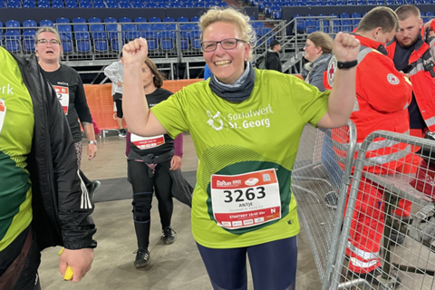 Eine Person mit Startnummer 3263 trägt ein grünes T-Shirt und hebt beide Arme, im Hintergrund stehen weitere Personen in Sportkleidung und Einsatzkräfte in roten Anzügen.