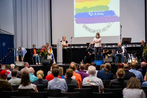 Mehrere Personen befinden sich auf einer Bühne vor einem sitzenden Publikum, im Hintergrund ist eine Leinwand mit Regenbogenfarben und dem Text „Gemeinsam für Vielfalt“ zu sehen.