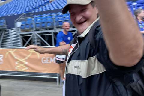 Eine Person mit Baseballkappe und Jacke hebt einen Arm, im Hintergrund sind weitere Personen und leere Sitzreihen in einem Stadion.